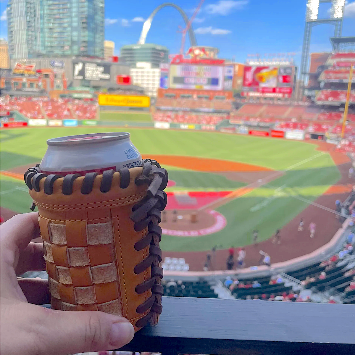 A tan leather baseball glove can cooler holding a silver can, held by a hand with a blurred baseball stadium background showing St. Louis Park and the Gateway Arch.

